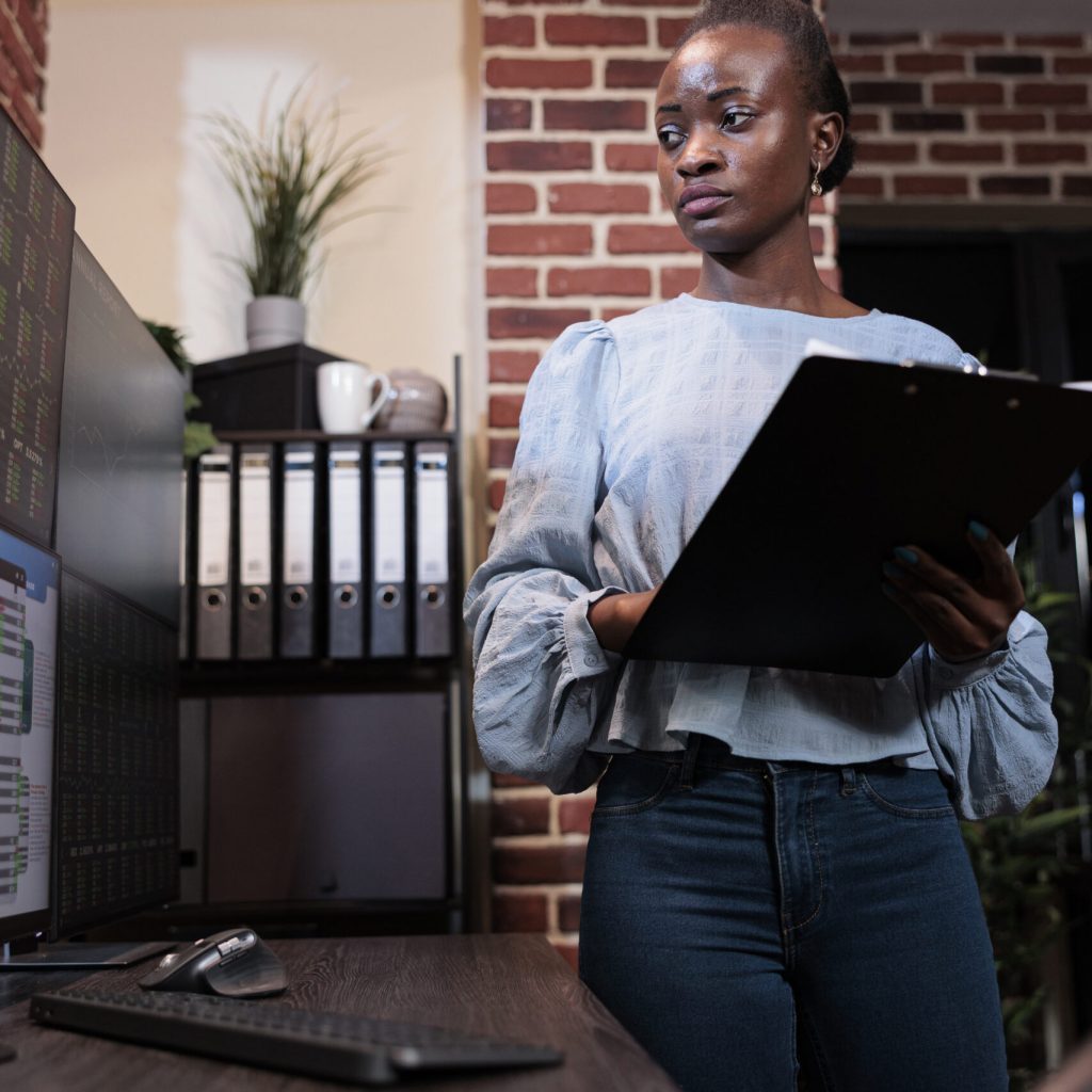 Investment company african american professional financial analyst with clipboard analyzing real time market data. Brokerage agency agent standing in front of multi monitor workstation with clipboard.